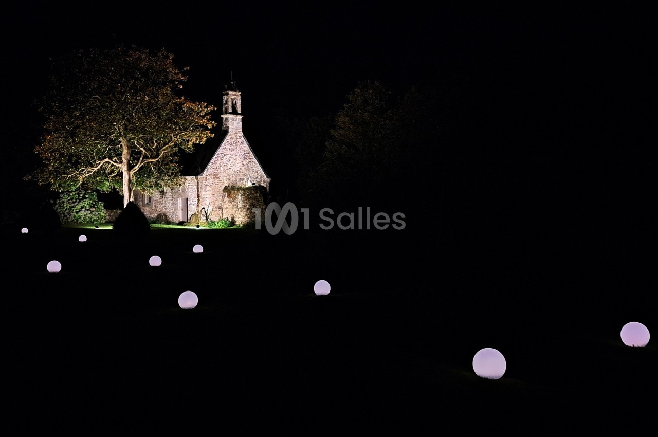 Petite chapelle en pierre éclairée la nuit, entourée d'arbres et de sphères lumineuses dans un jardin sombre.