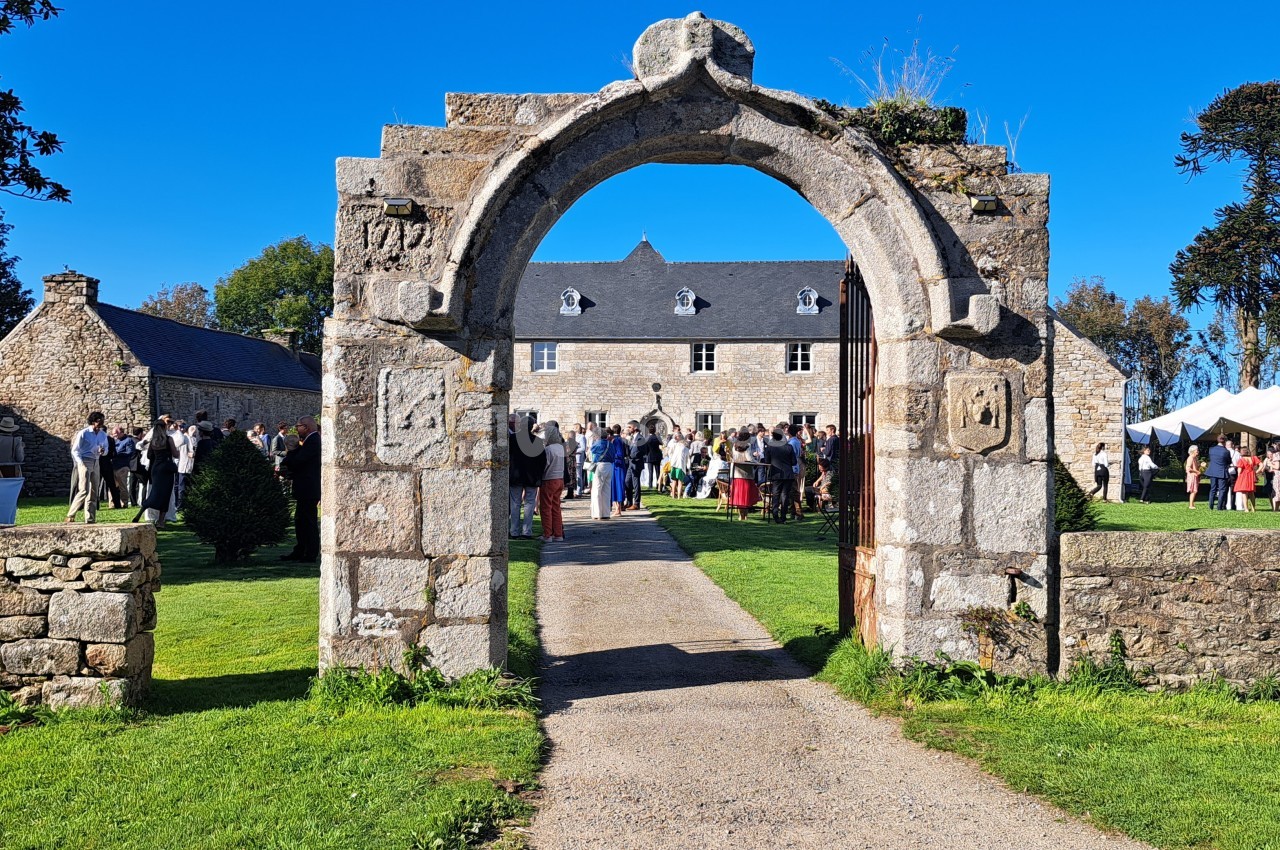 Portail en pierre donnant sur une cour avec des bâtiments anciens, des invités et des tentes blanches en arrière-plan.