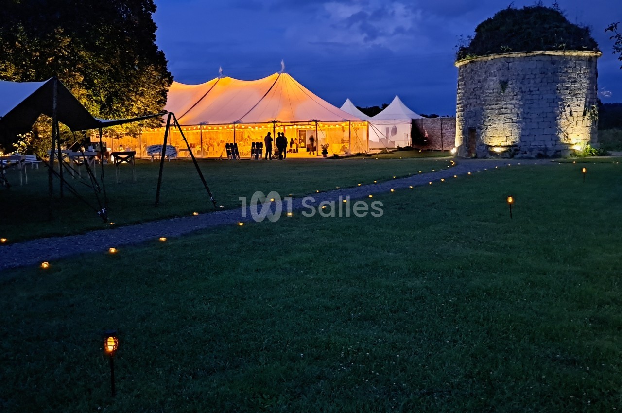 Tente éclairée pour un événement nocturne, entourée de pelouse et d'un ancien bâtiment en pierre.