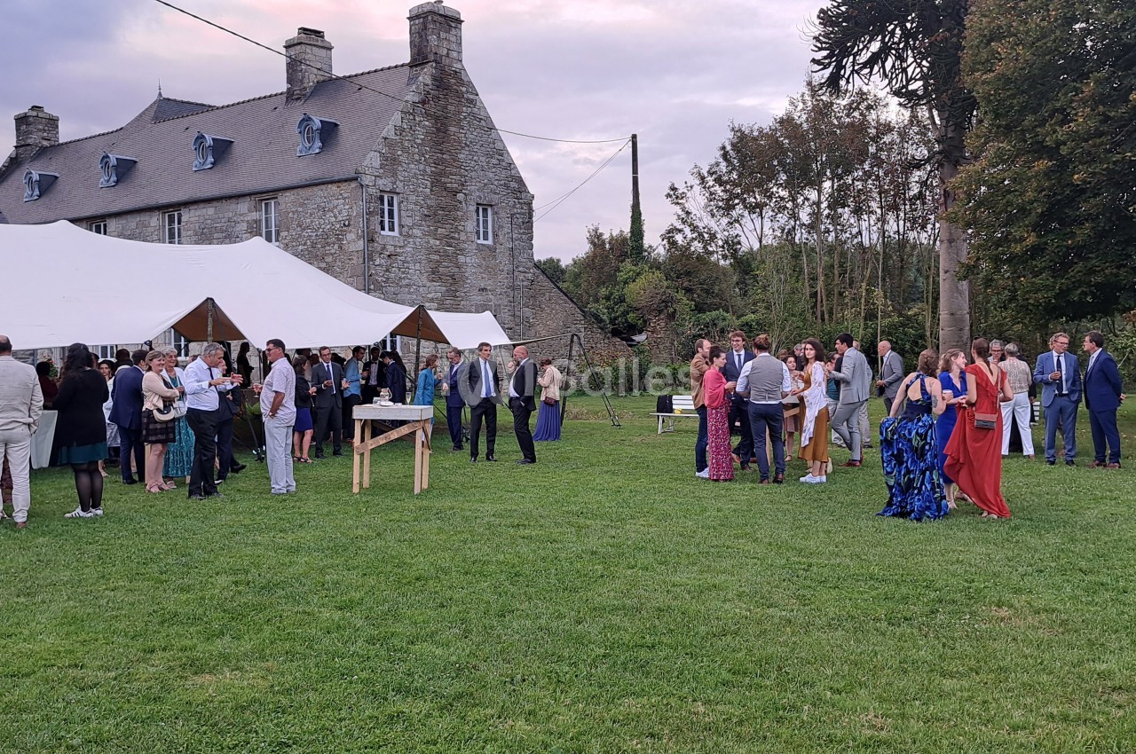 Groupe de personnes rassemblées dans un jardin devant un bâtiment en pierre, avec des tentes blanches en arrière-plan.