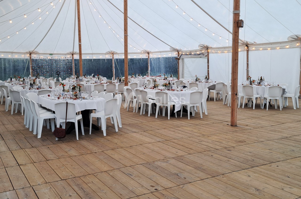 Salle sous chapiteau avec tables dressées, nappes blanches et chaises en plastique, éclairée par des guirlandes lumineuses.