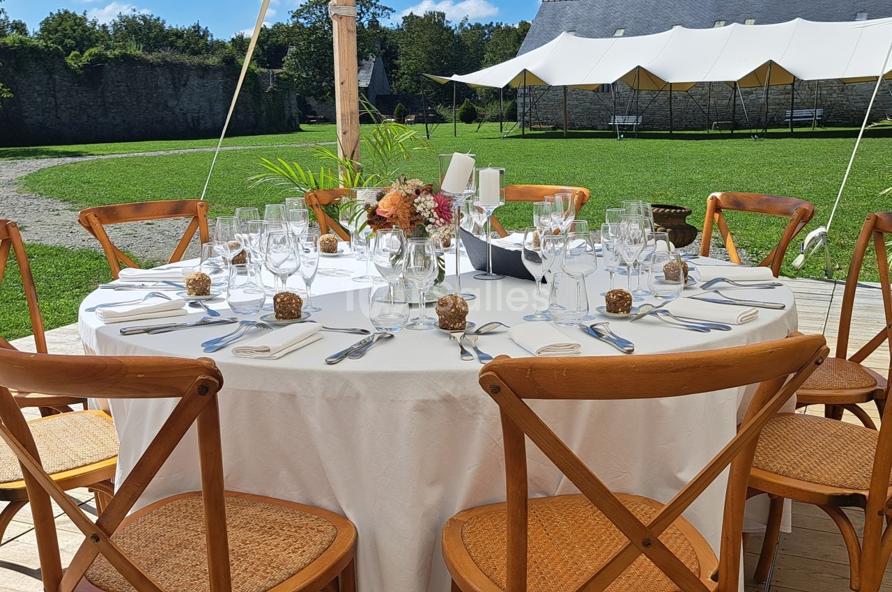 Table ronde dressée pour un repas en extérieur, avec nappes blanches, chaises en bois et décoration florale au centre.