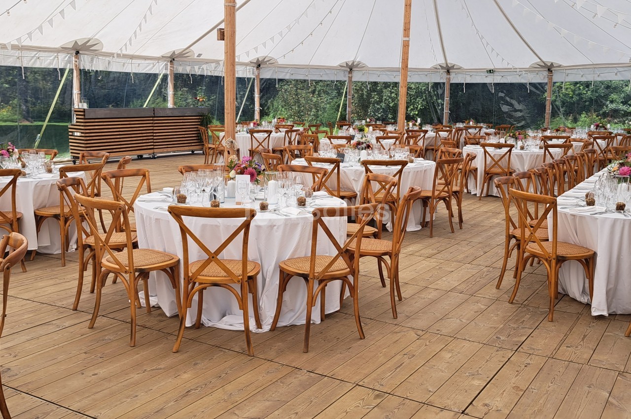 Salle de réception en plein air sous une tente, avec tables rondes dressées et chaises en bois sur un plancher en bois.