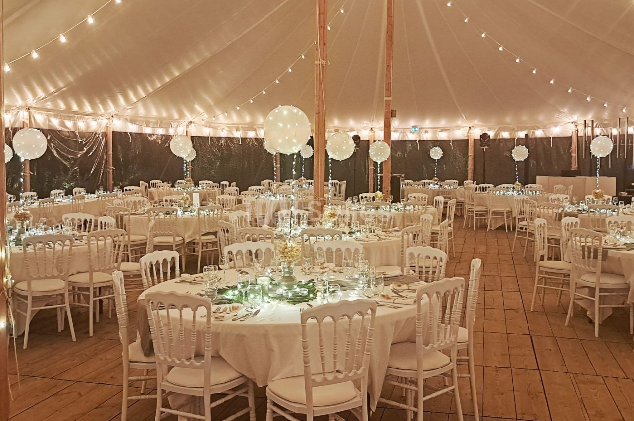 Salle de réception sous une tente éclairée, avec des tables rondes décorées et des chaises blanches alignées.