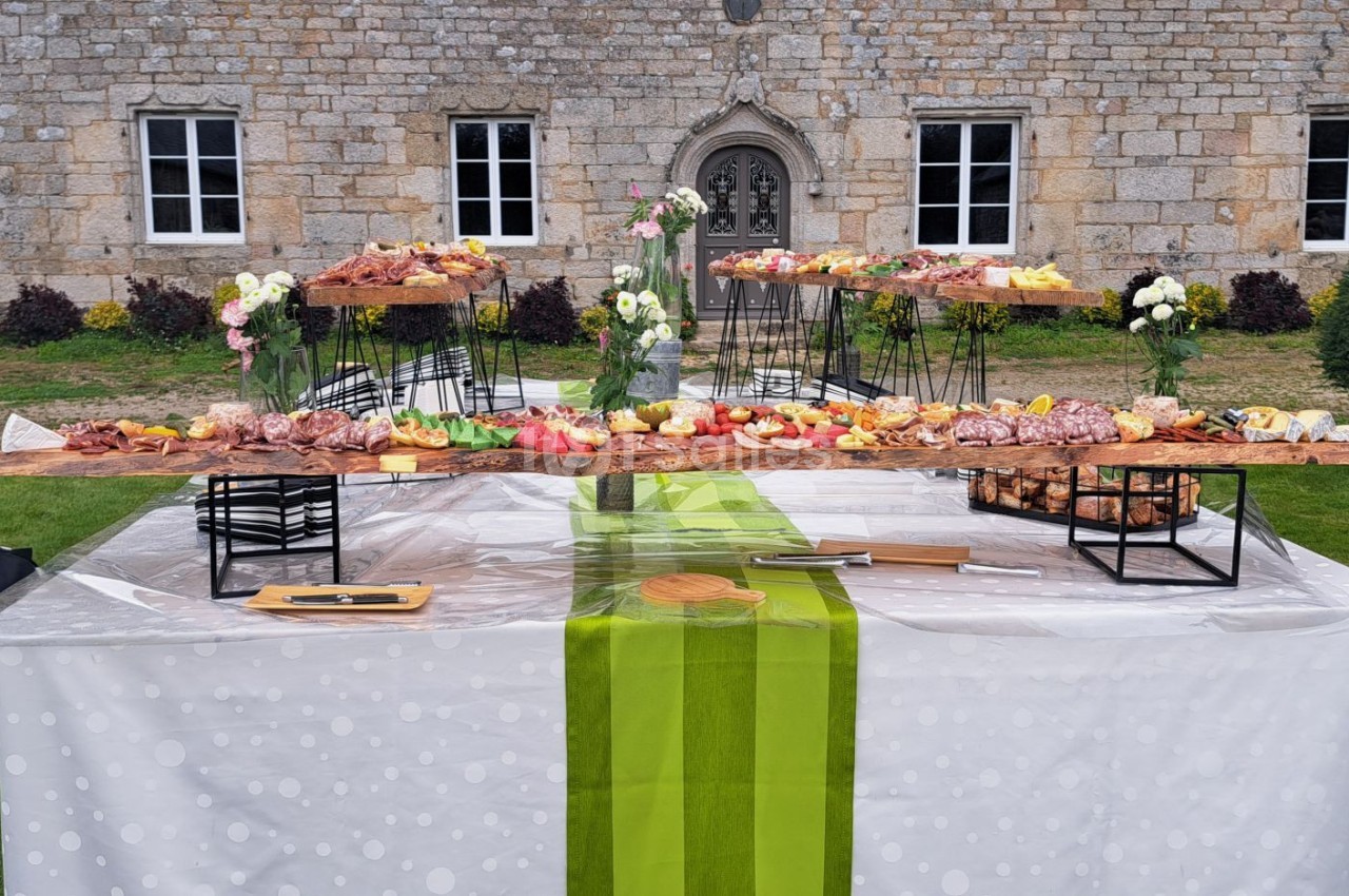 Table garnie de charcuteries, fromages et fruits, disposée sur une pelouse devant un bâtiment en pierre.