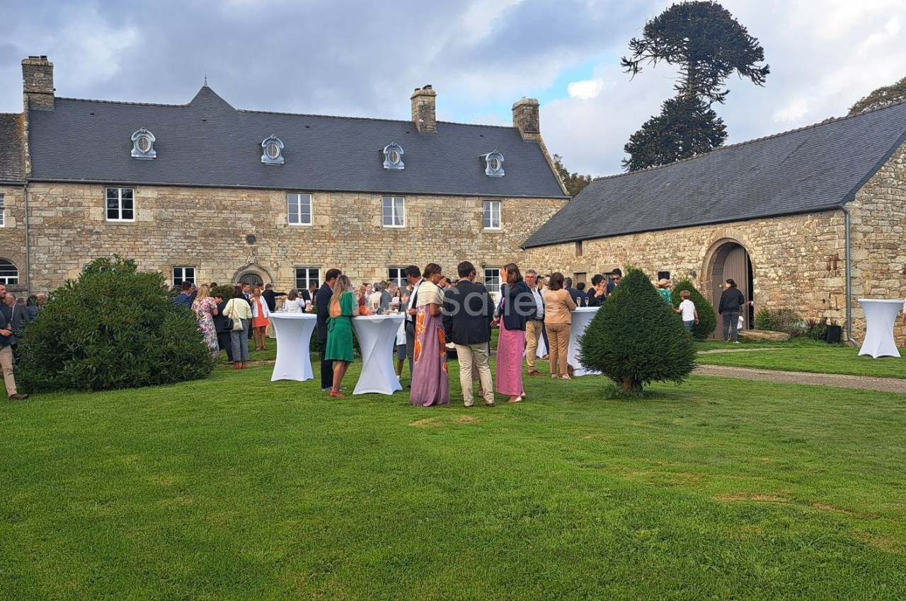Groupe de personnes rassemblées dans un jardin devant un bâtiment en pierre, lors d'un événement en plein air.