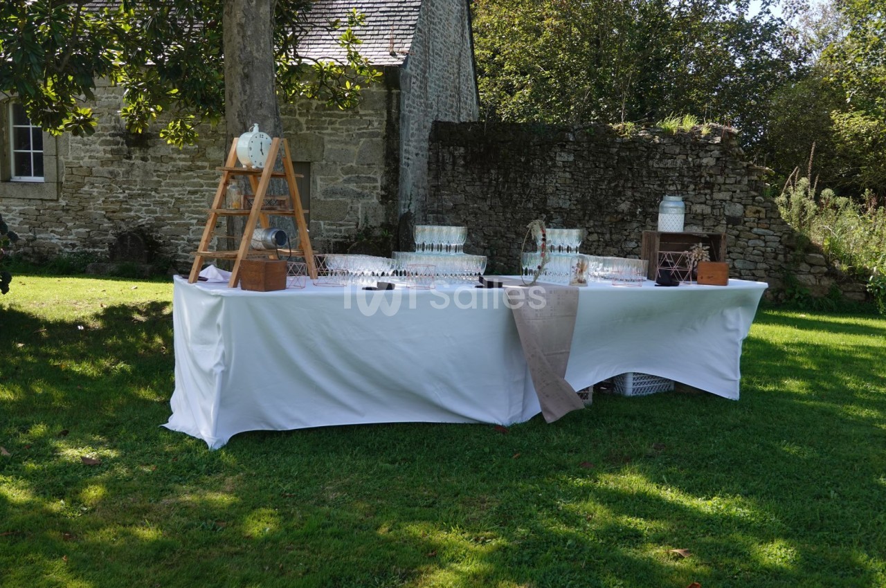 Table dressée en extérieur avec nappes blanches, verres alignés et décorations rustiques près d'un mur en pierre.