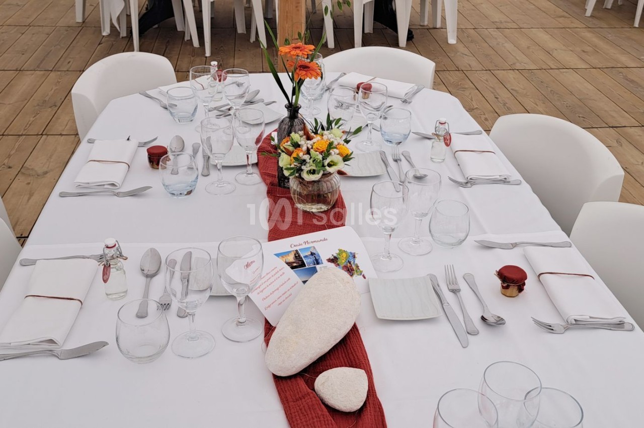Table dressée avec nappe blanche, chemin de table rouge, vaisselle, fleurs et décorations en pierre sur une terrasse en bois.
