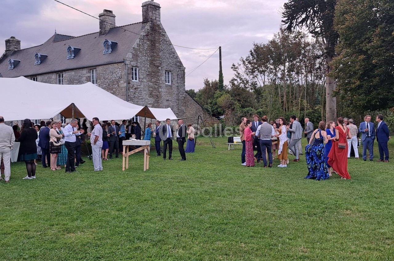 Groupe de personnes rassemblées dans un jardin devant une grande maison en pierre et des tentes blanches.