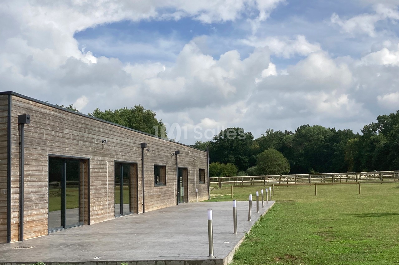 Bâtiment en bois avec une terrasse en béton, entouré de pelouses et de clôtures dans un environnement rural.