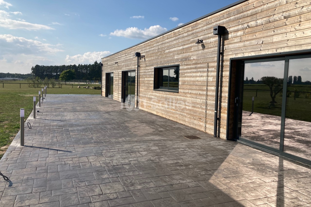 Bâtiment moderne en bois avec de grandes baies vitrées, entouré d'une terrasse en béton et d'un espace vert.