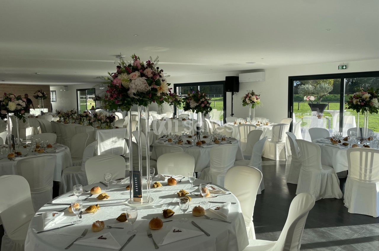 Salle de réception lumineuse avec tables décorées de nappes blanches, centres de table fleuris et vaisselle élégante.