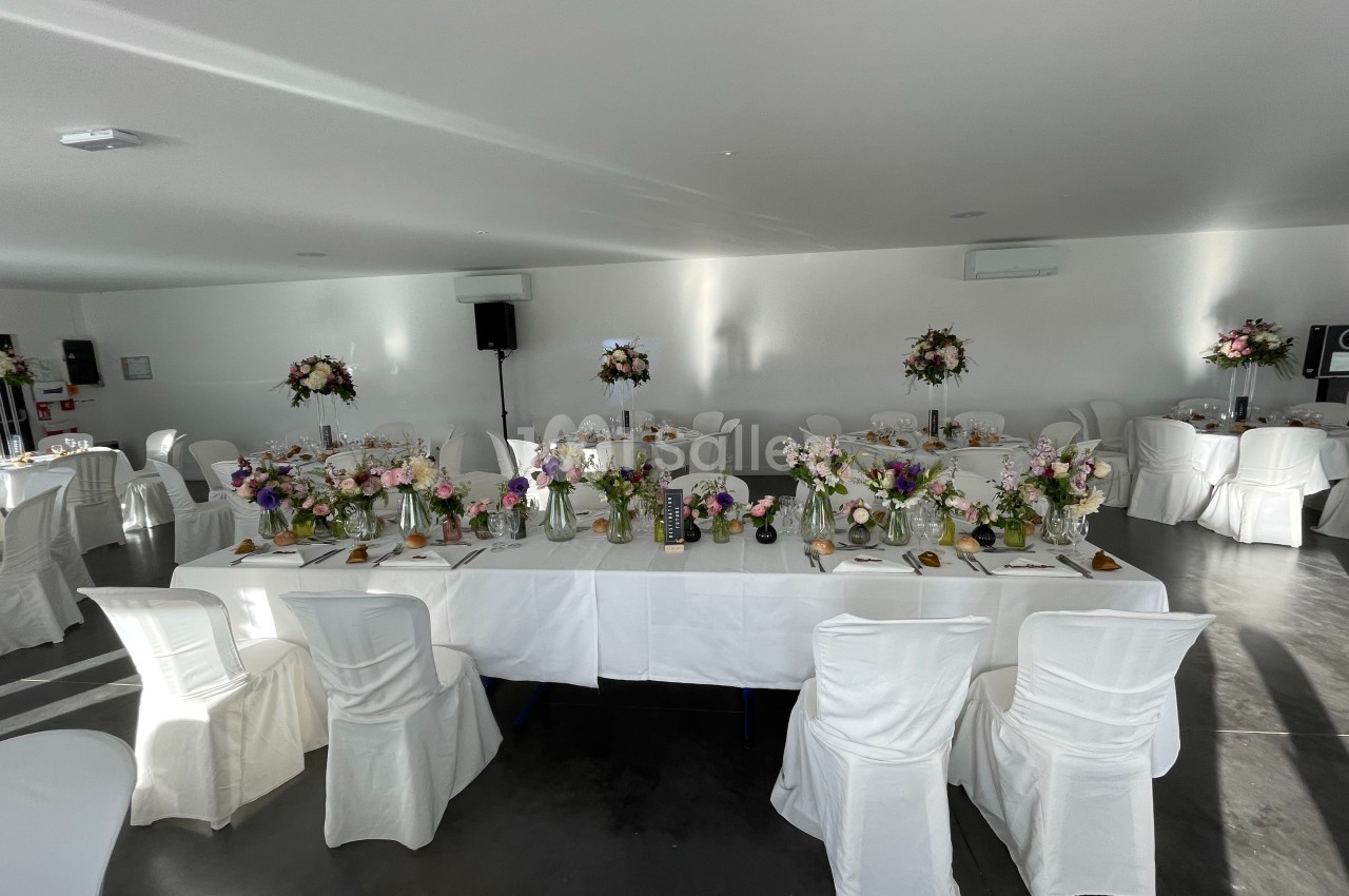 Salle de réception décorée pour un événement, avec des tables ornées de fleurs et des chaises recouvertes de housses…