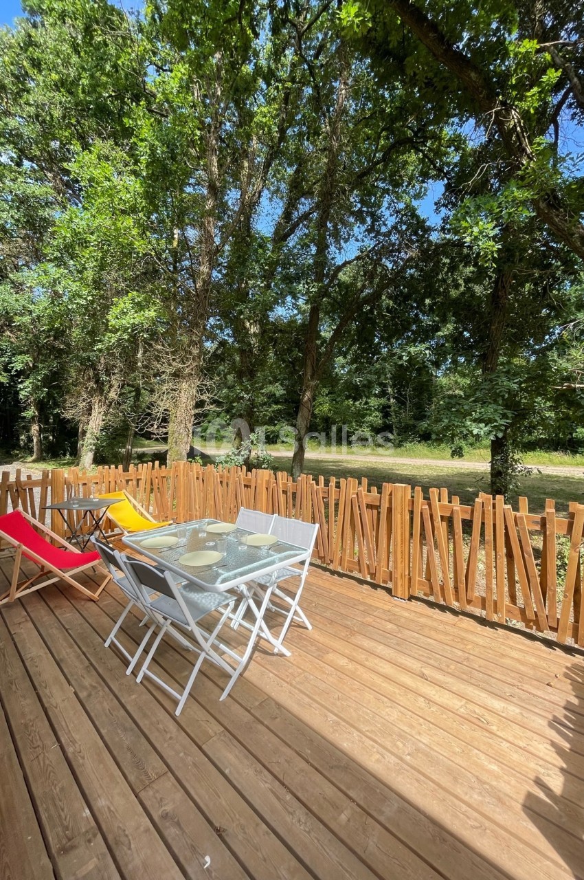 Terrasse en bois avec table, chaises blanches et transats colorés, entourée d'une clôture et bordée d'arbres.
