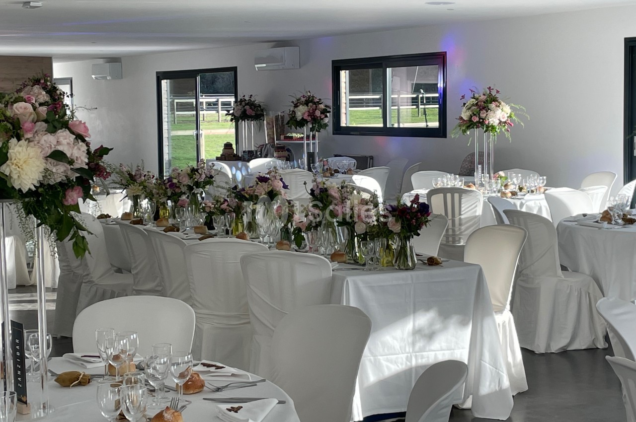Salle de réception décorée pour un mariage, avec tables dressées, nappes blanches et arrangements floraux élégants.