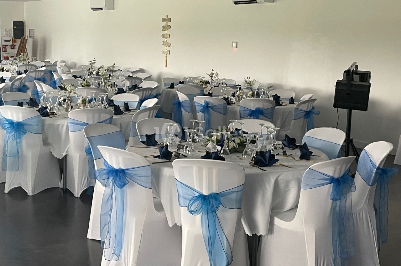 Salle de réception décorée avec des tables rondes, nappes blanches et chaises ornées de nœuds en tissu bleu.