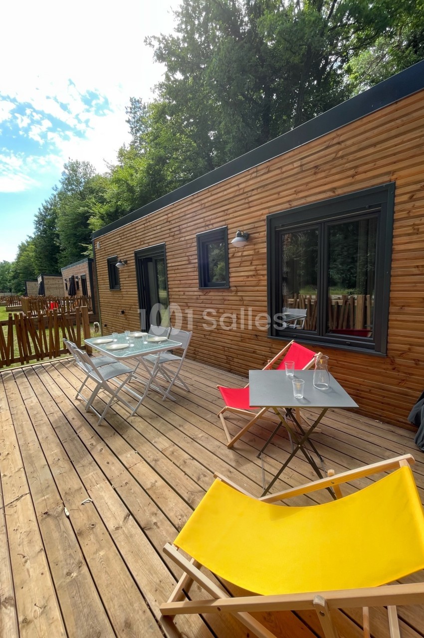 Terrasse en bois avec table, chaises et transats colorés, devant un logement en bois entouré de verdure.