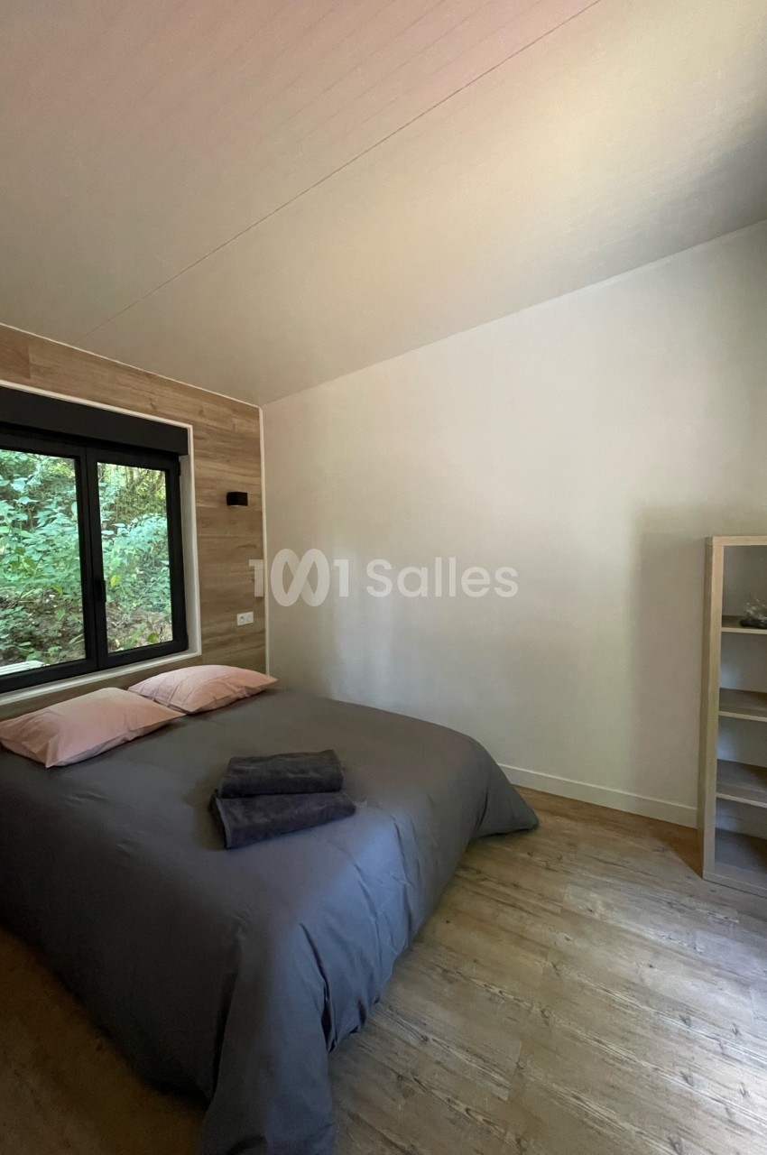 Chambre avec lit double, draps gris, oreillers roses, étagère en bois et fenêtre donnant sur un paysage boisé.