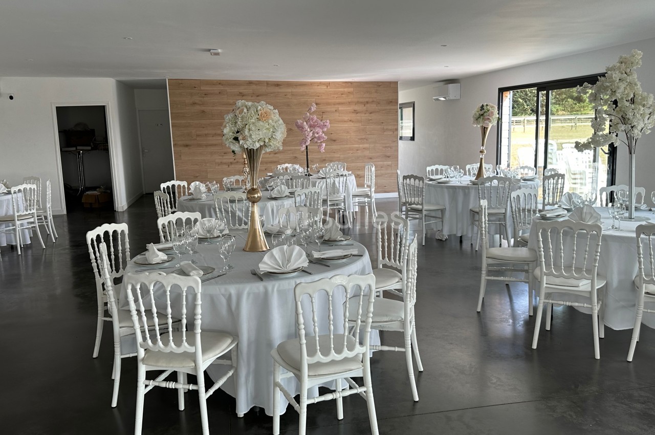 Salle de réception lumineuse avec tables rondes dressées, nappes blanches et chaises blanches, décor floral élégant.