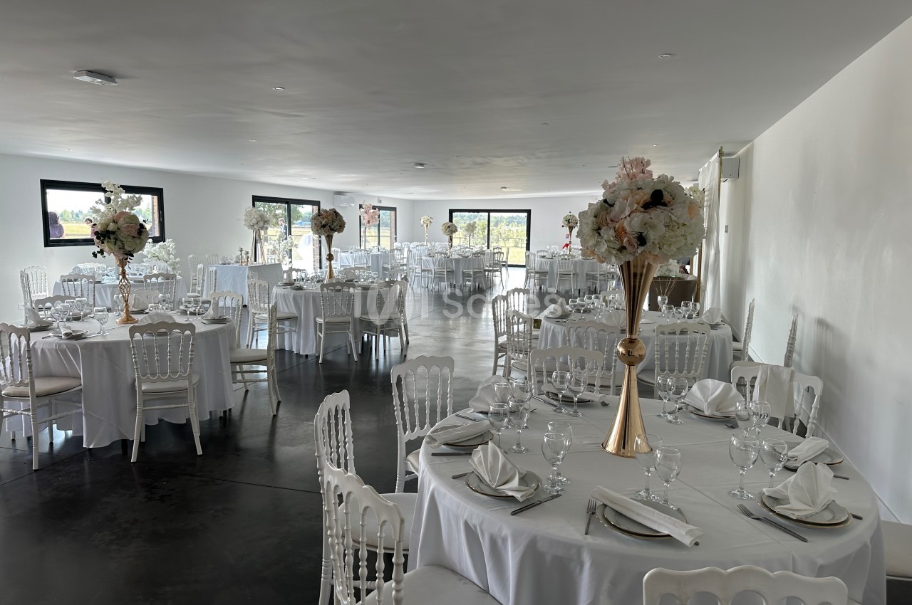 Salle de réception lumineuse avec tables rondes dressées, nappes blanches et centres de table fleuris.