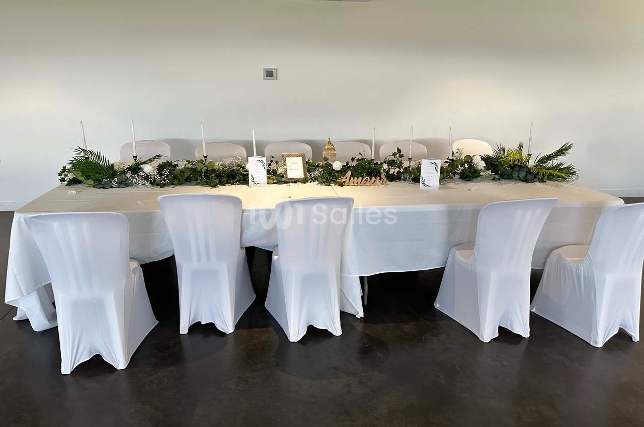 Table décorée avec une nappe blanche, des chaises houssées, un centre de table végétal et des bougies dans une salle…