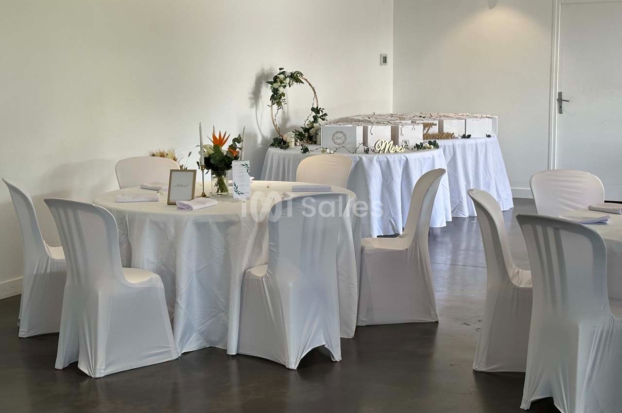 Salle décorée pour un événement avec tables rondes nappées de blanc, chaises assorties et buffet en arrière-plan.