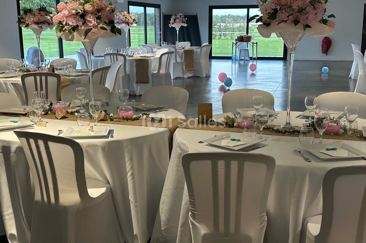 Salle de réception décorée pour un mariage avec tables rondes, nappes blanches, centres de table fleuris et baies vitrées.