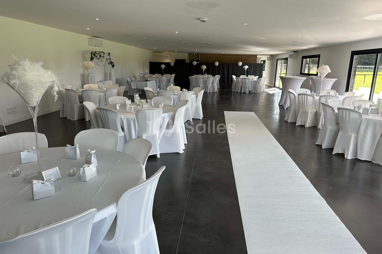 Salle de réception décorée avec des tables rondes, nappes blanches, chaises houssées et allée centrale blanche.