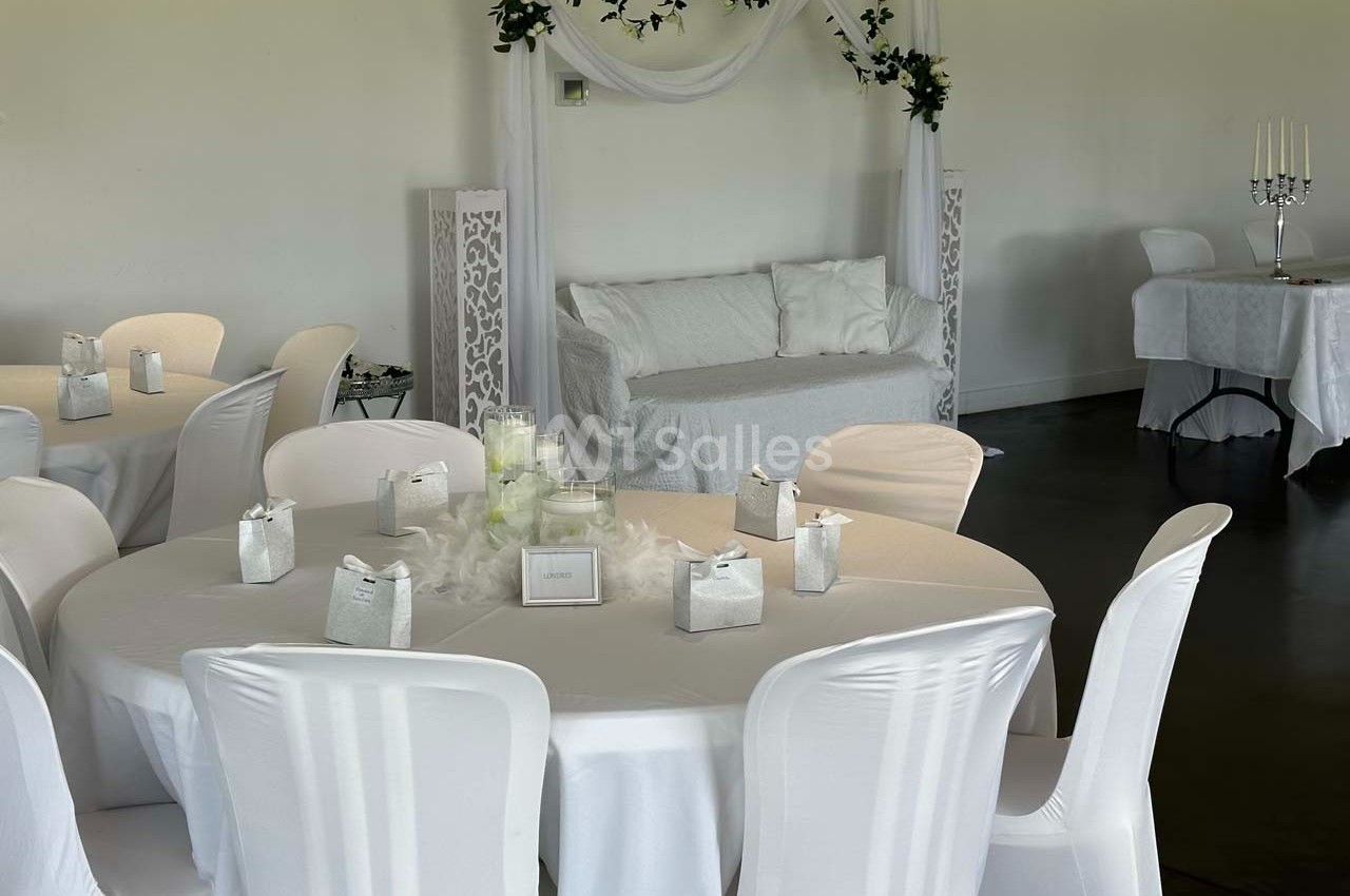Salle décorée pour un événement avec des tables rondes couvertes de nappes blanches et une arche ornée de voilages.