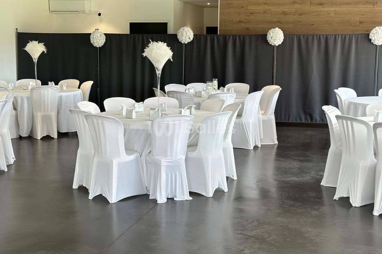 Salle de réception avec des tables rondes dressées, nappes blanches et décorations florales blanches sur pied.