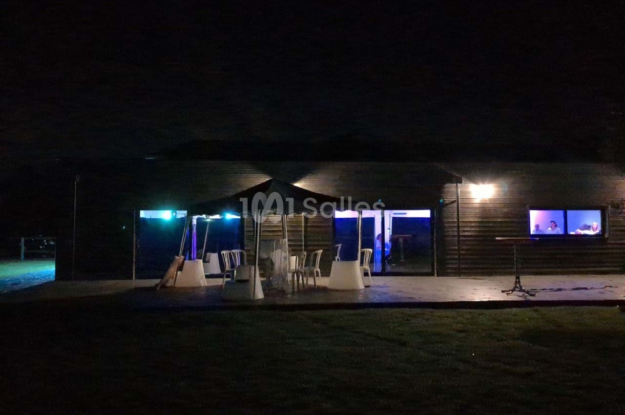 Terrasse extérieure sombre avec des chaises et une table sous un parasol, éclairée par des lumières bleues et jaunes.