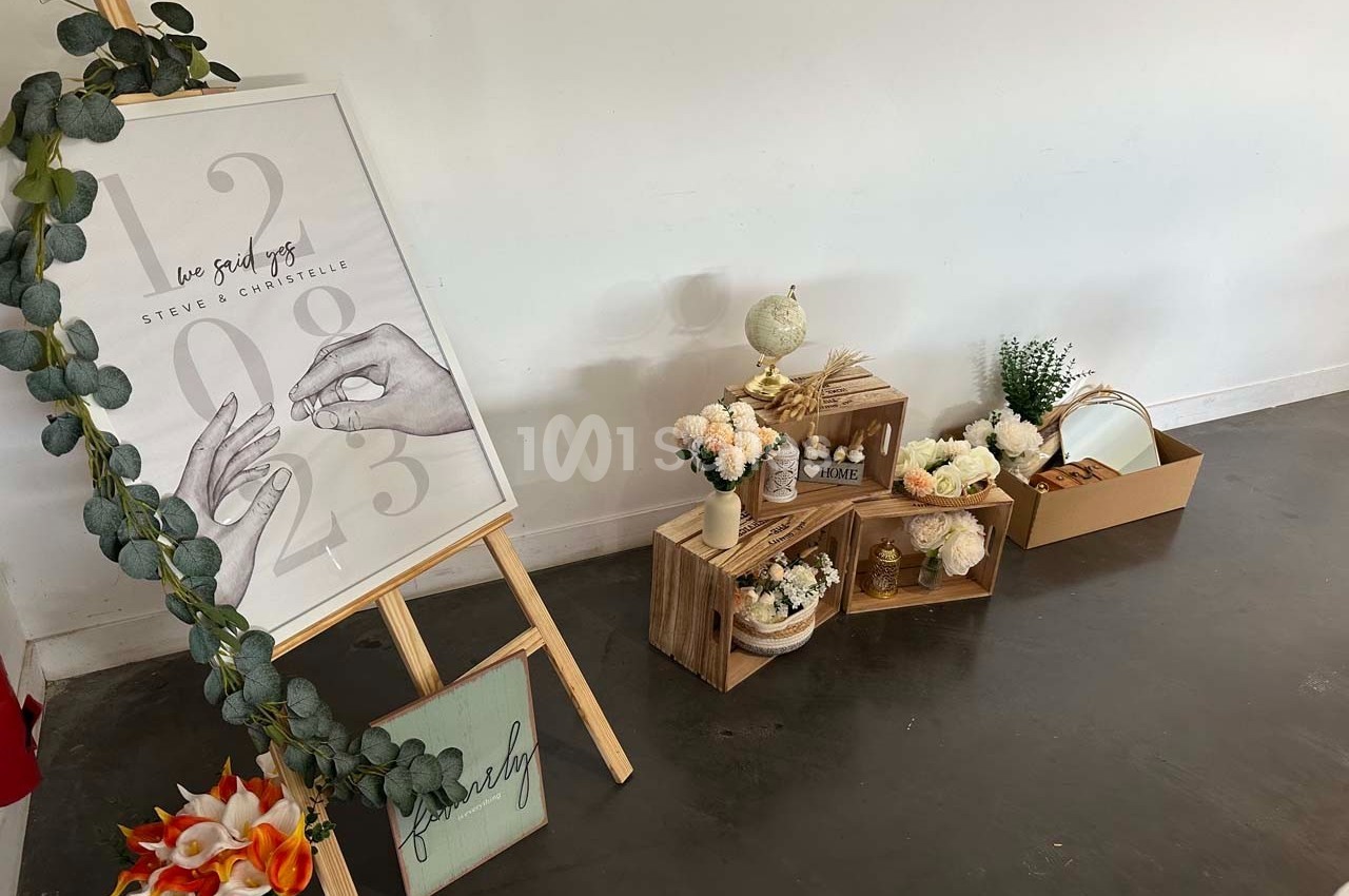 Décoration de mariage avec un chevalet illustré, des fleurs, des caisses en bois et des accessoires sur un sol sombre.