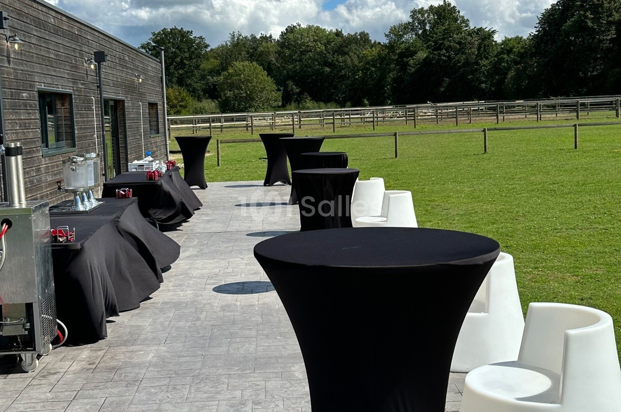 Tables hautes noires et chaises blanches disposées sur une terrasse ensoleillée, avec vue sur un grand espace vert.