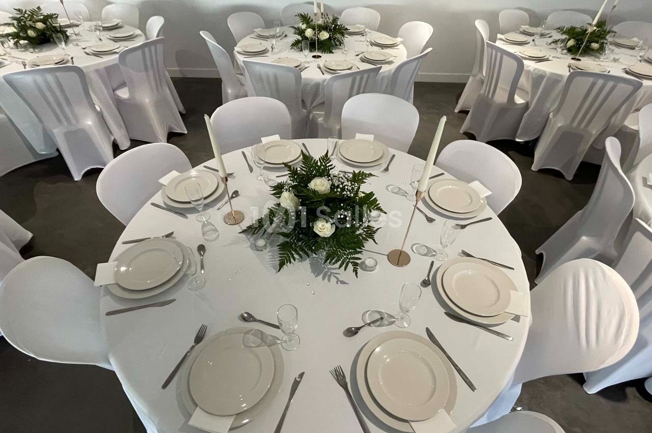 Tables rondes dressées avec nappes blanches, vaisselle élégante et centres de table composés de fleurs et de feuillage.