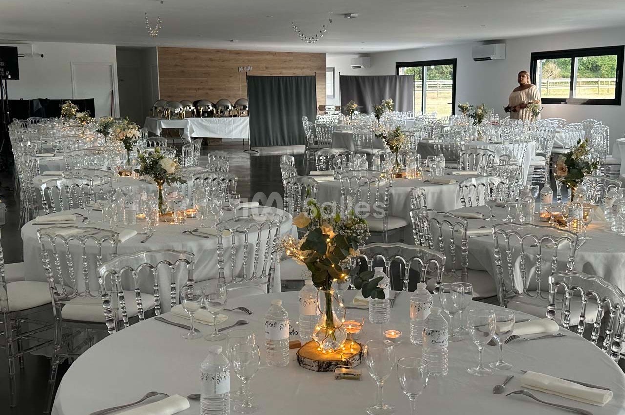 Salle de réception décorée avec des tables rondes, nappes blanches, chaises transparentes et centres de table fleuris.