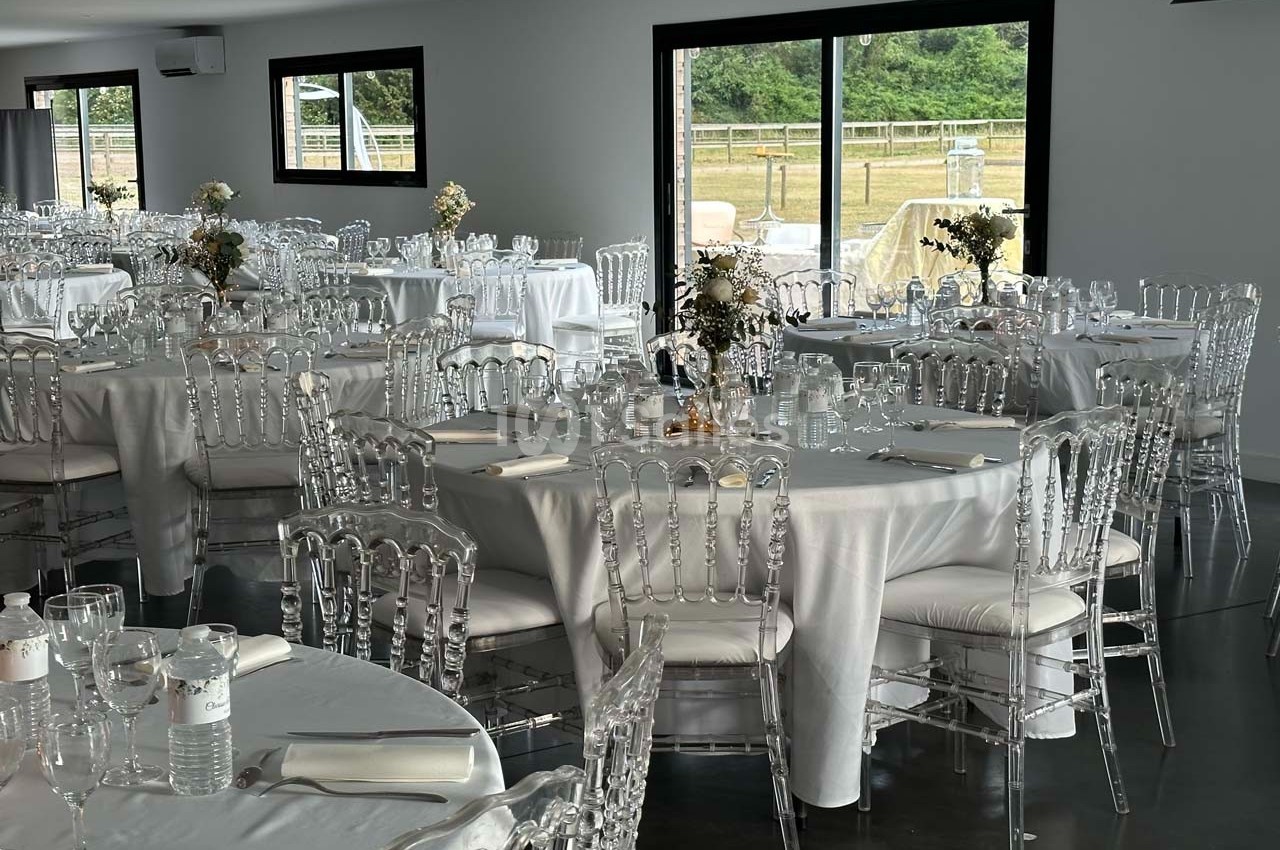 Salle de réception lumineuse avec des tables rondes dressées de nappes blanches et des chaises transparentes.