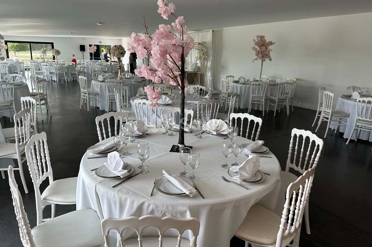 Salle de réception avec tables rondes dressées, nappes blanches et centres de table décorés de branches fleuries roses.