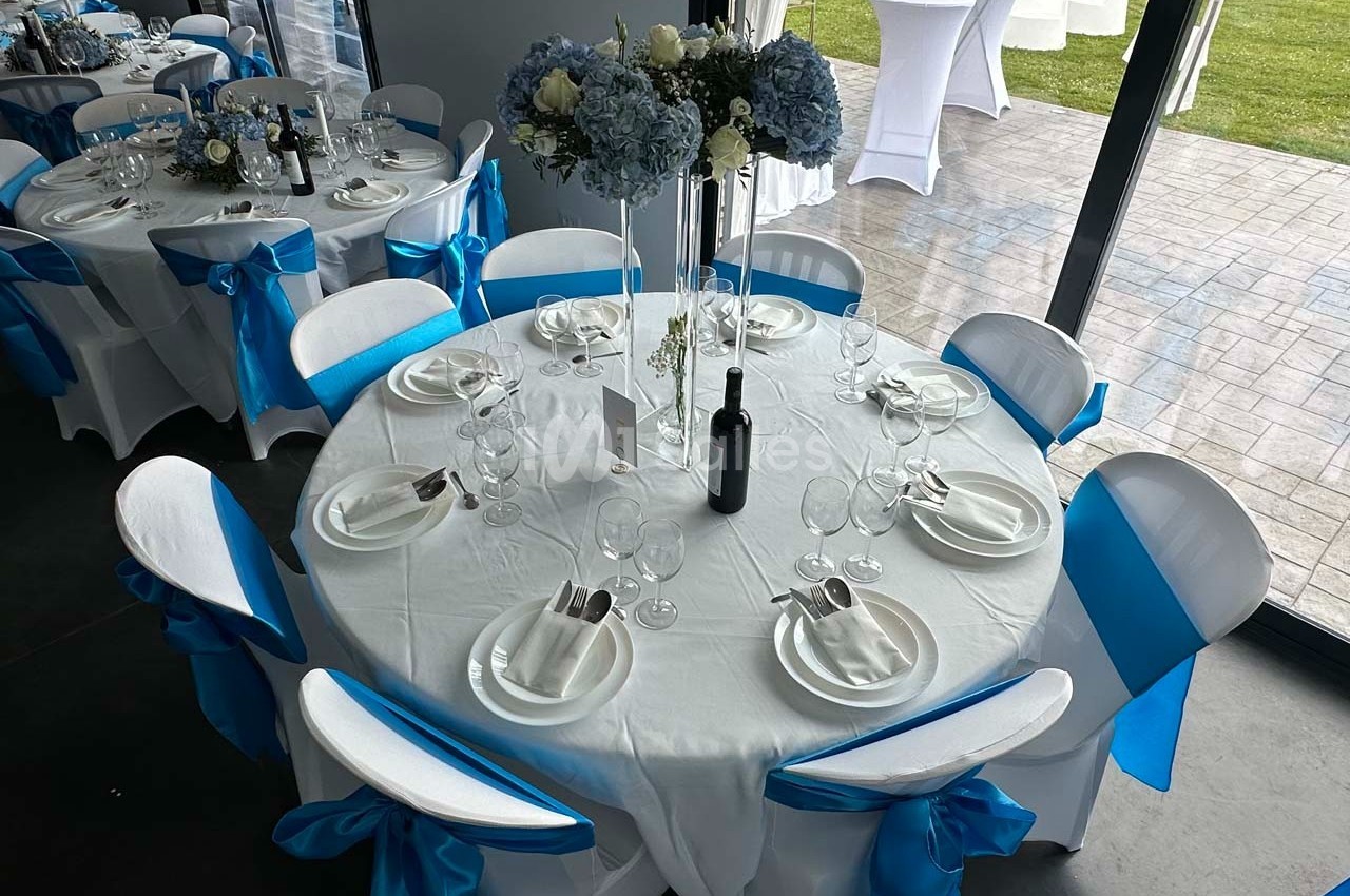 Tables rondes décorées de nappes blanches et de nœuds bleus, avec des centres de table floraux et des couverts disposés.