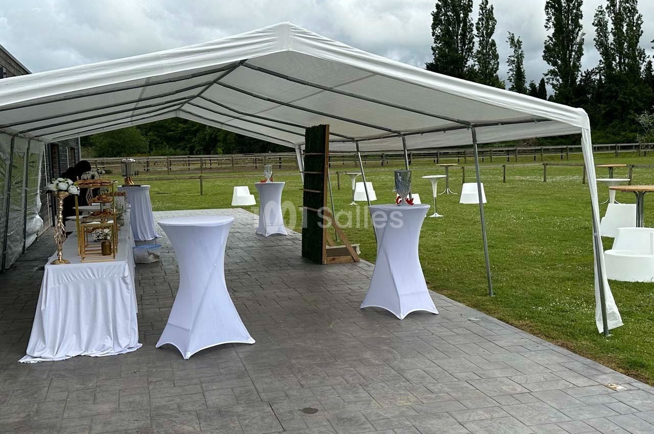 Tente blanche installée sur une terrasse pavée, avec des tables hautes habillées de nappes blanches et un espace vert en…