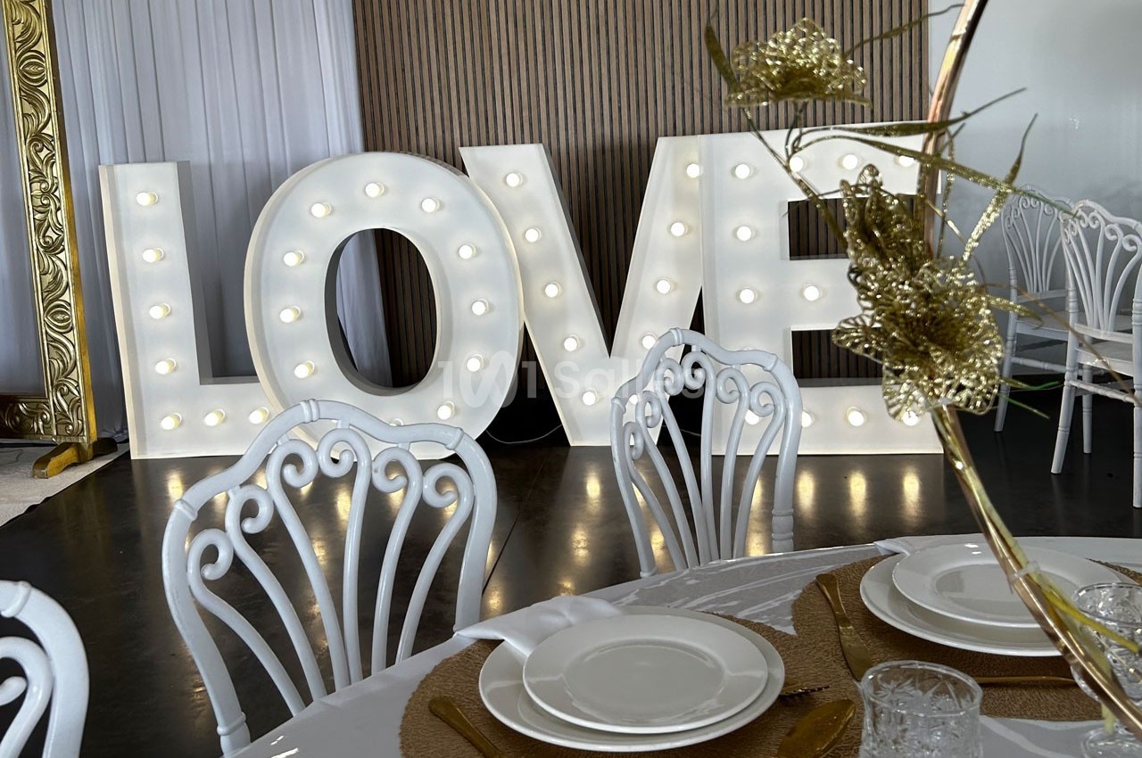 Lettres lumineuses formant le mot ’LOVE’ décorant une salle avec des chaises blanches et une table dressée.