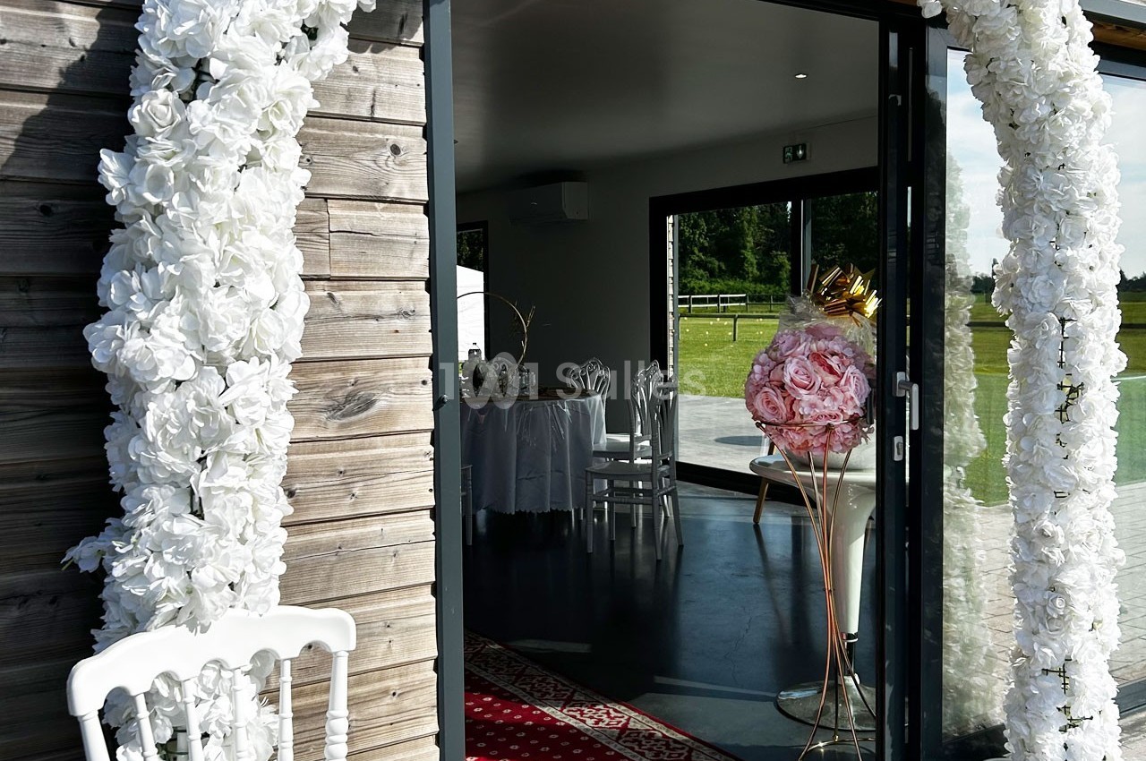 Entrée décorée de fleurs blanches menant à une salle de réception lumineuse avec des chaises et une table dressée.
