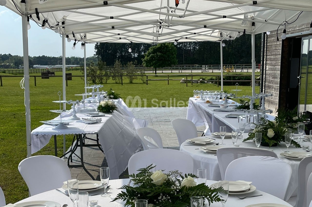 Tables dressées avec nappes blanches et décorations florales sous des tonnelles, dans un espace extérieur verdoyant.