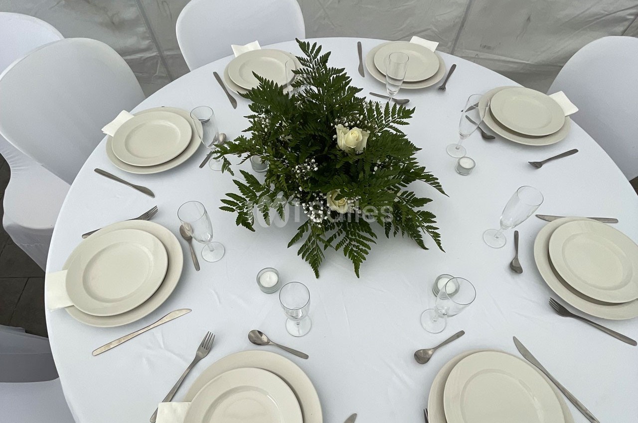 Table ronde dressée avec une nappe blanche, vaisselle blanche, couverts en métal et un centre de table floral.