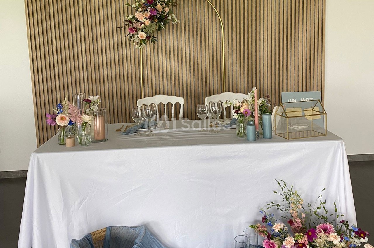 Table décorée pour un mariage avec nappes blanches, fleurs colorées, verrerie et arche florale en arrière-plan.