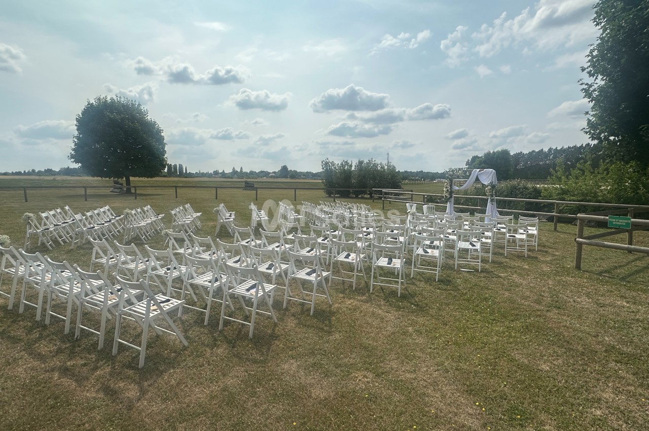 Chaises blanches disposées en rangées sur une pelouse, face à une arche décorée, dans un cadre extérieur champêtre.