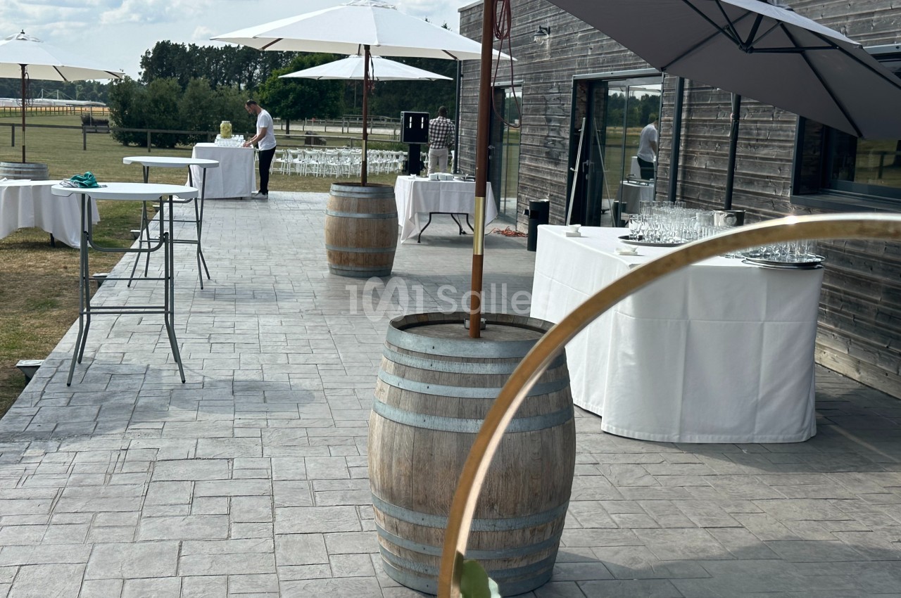 Terrasse extérieure avec tables dressées, parasols et tonneaux en bois, prête pour un événement en plein air.