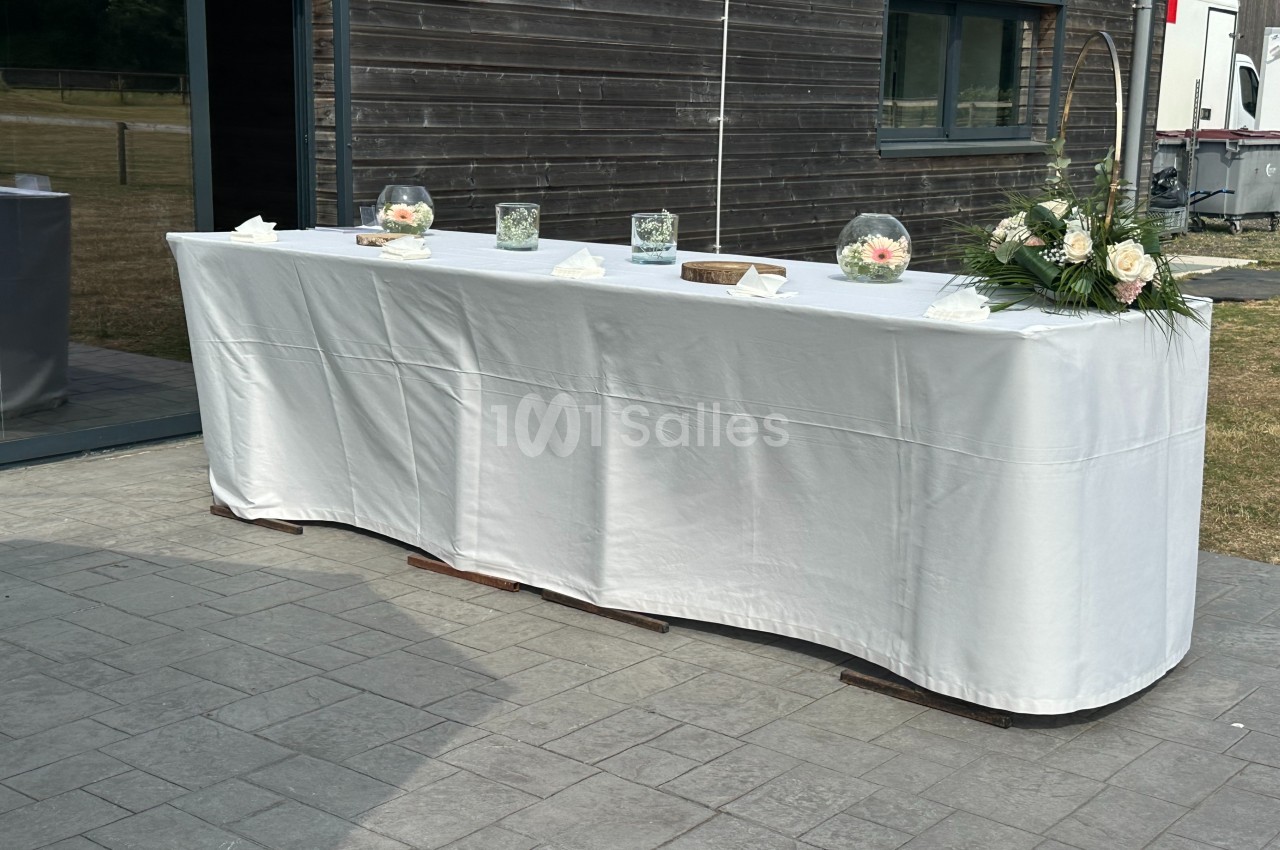 Table recouverte d'une nappe blanche, décorée de fleurs et de vases, placée devant un bâtiment en bois.