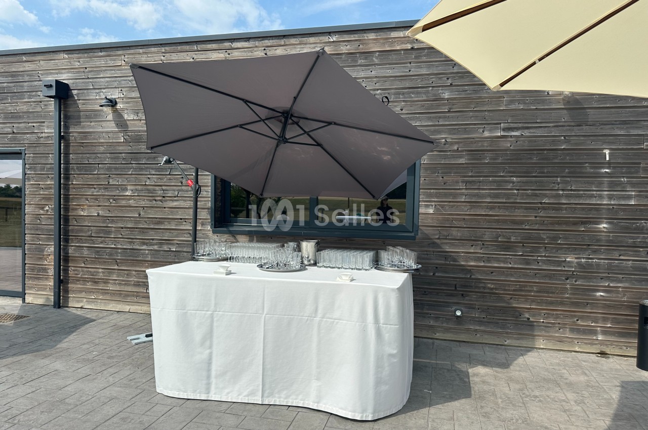 Table recouverte d'une nappe blanche avec verres et seaux à glace, abritée par des parasols devant un mur en bois.