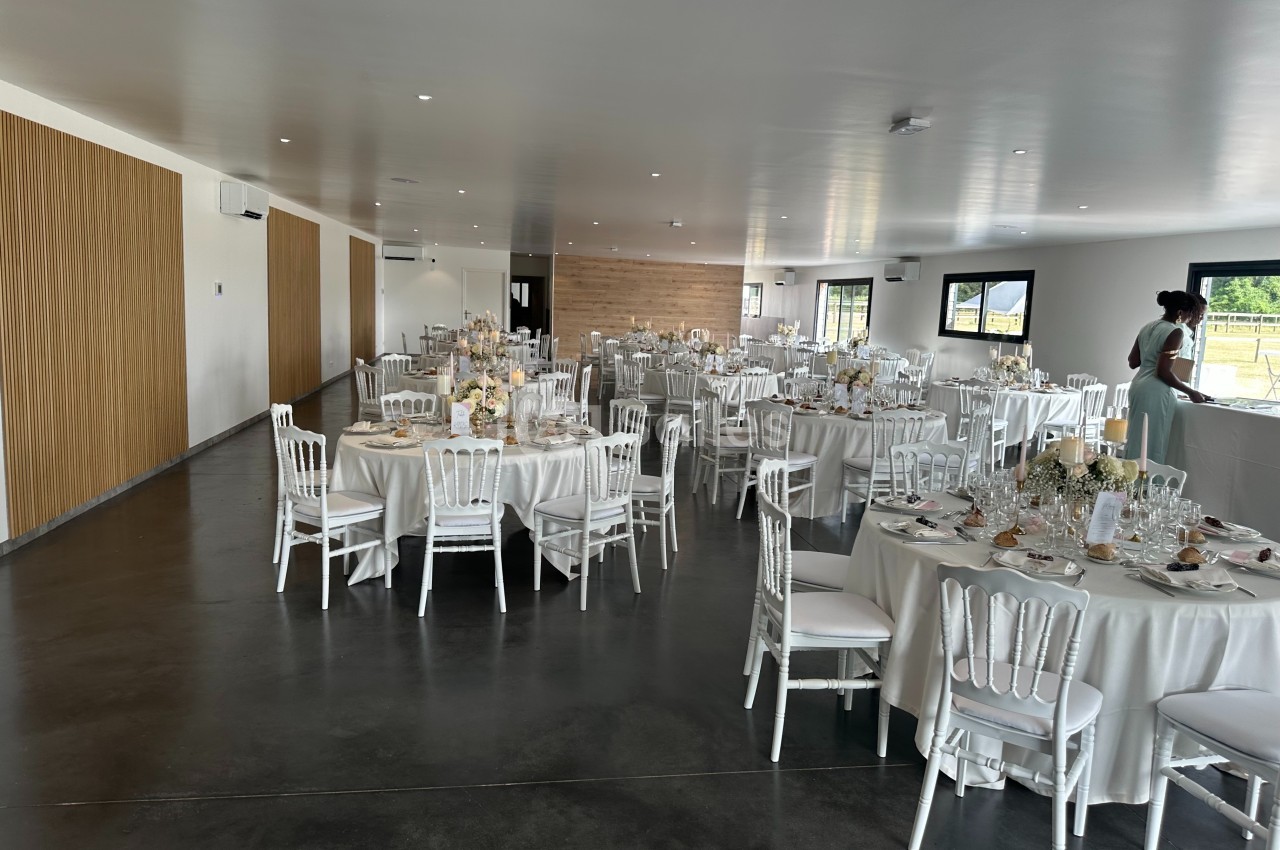 Salle de réception lumineuse avec tables rondes dressées, chaises blanches et décorations florales élégantes.