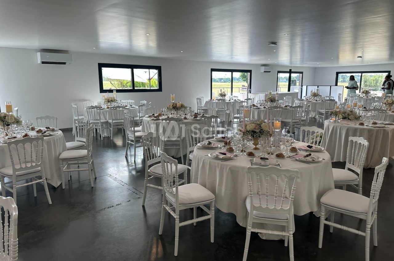 Salle de réception lumineuse avec tables rondes décorées de nappes blanches, fleurs et vaisselle élégante.